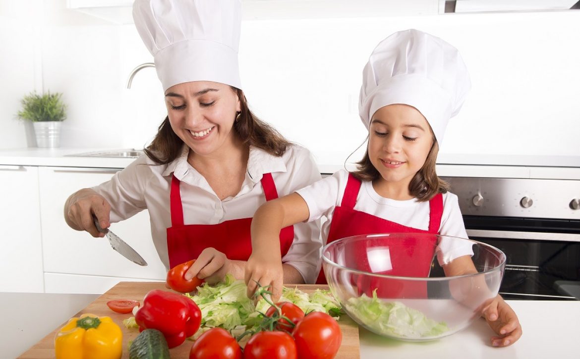 young mother and little daughter at house kitchen preparing salad for lunch wearing apron and cook hat